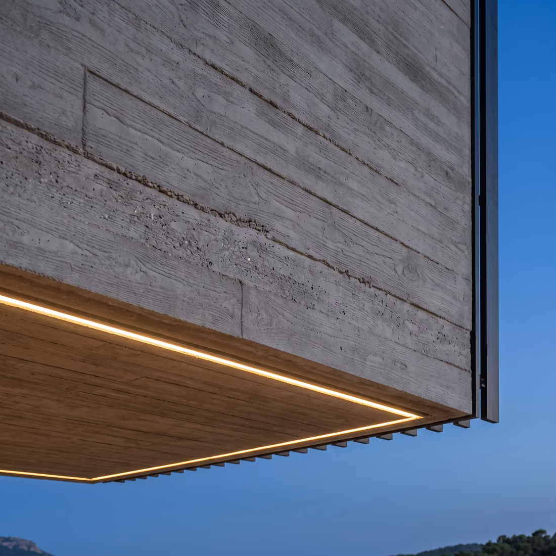 Sous-face du porte-à-faux en béton banché, veinée par les planches du coffrage et soulignée d'un filet de lumière.